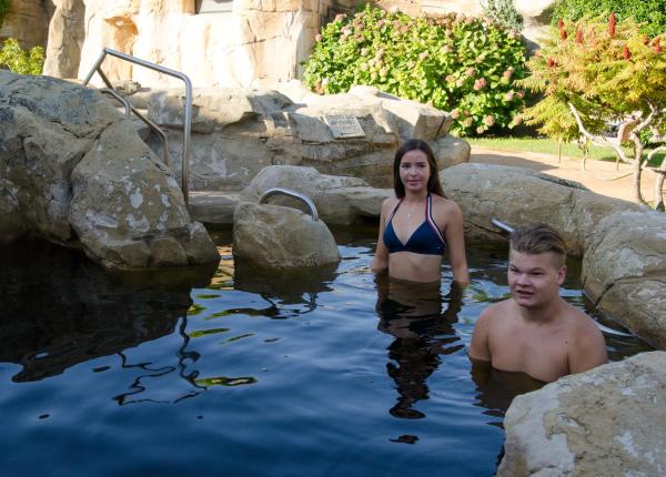Twee mensen ontspannen in een natuurlijke warmwaterbron, omgeven door rotsen en planten.